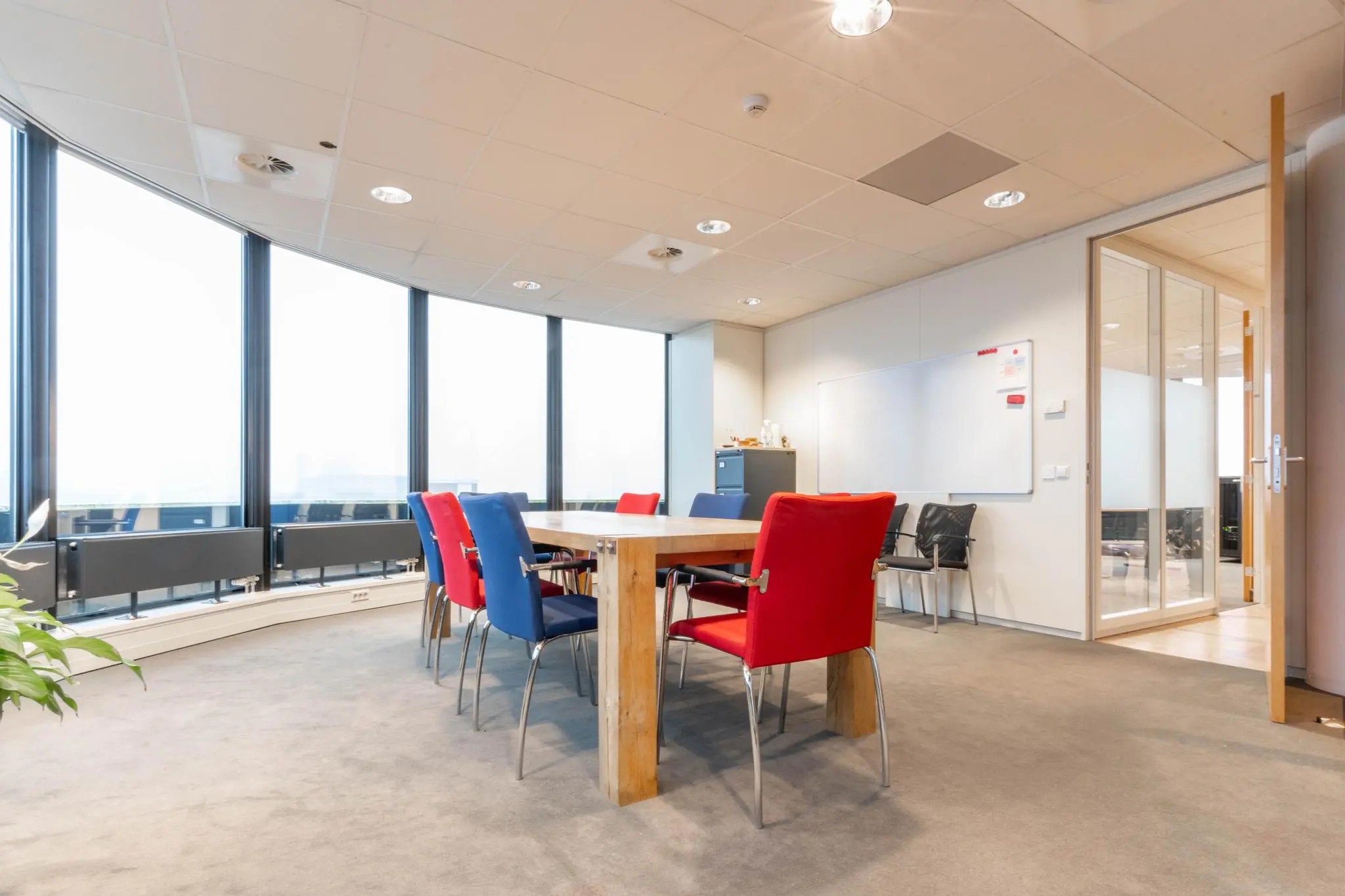 Vergaderruimte met kleurrijke stoelen rond een houten tafel en grote ramen aan de 1e Hogeweg.